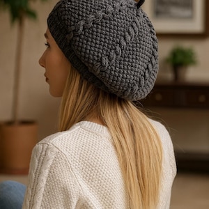 Könnte beinhalten: Ein grauer Strickbarett mit strukturiertem Muster und kleinem Stieldetail. Das Barett wird mit einem weißen Pullover getragen. Der Hut ist ein stilvolles Accessoire für kaltes Wetter.