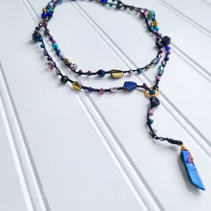 May include: A long, layered necklace featuring a variety of colourful beads, including gold, blue, purple, and turquoise. The necklace has a long, dangling pendant made of a blue crystal.