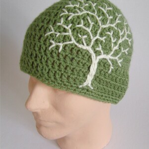May include: A green crocheted beanie featuring a white tree design. The tree is embroidered on the front of the hat. The hat is made of a soft, textured yarn. The beanie is shown on a mannequin head.