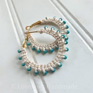 May include: A pair of handmade cream-colored macrame hoop earrings with gold-colored clasps. The earrings are adorned with small, turquoise-colored beads. The earrings are displayed on a white surface.