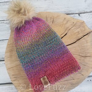 May include: A colorful knitted beanie with a faux fur pom pom and a leather tag with a logo. The beanie is made in a rainbow color scheme with shades of pink, purple, blue, green, and yellow.