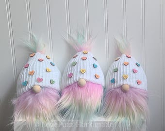 Könnte beinhalten: Drei Gnome mit weißen Strickmützen, die mit bunten Herzchen aus Süßigkeiten verziert sind. Die Gnome haben langes, rosa und blaues Ombre-Fell.