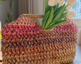 May include: A handwoven tote bag in shades of gold, pink, and brown, with a woven handle. The bag is filled with white tulips. The bag's texture is visible, and the colors transition from pink to gold.