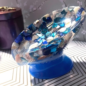 Può includere: Un portasapone in resina blu e bianca con un design decorativo di conchiglie e pezzi di vetro blu incassati. Il portasapone ha la forma di una ciotola e ha una base blu.