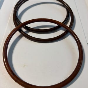 May include: Two circular, dark brown, smooth-surfaced bag handles. The rings are constructed from a solid, glossy material with a uniform, rounded profile. They are positioned overlapping each other, showcasing a consistent deep brown color throughout.