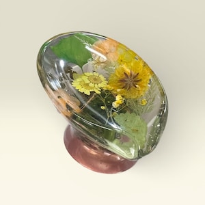 May include: A clear, oval-shaped resin paperweight with dried yellow and orange flowers and green leaves encased inside. The paperweight sits on a copper-colored base.