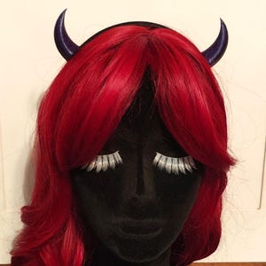 Può includere: Un busto di manichino nero con una parrucca rossa e corna diaboliche viola. La parrucca ha capelli lunghi e mossi. Le ciglia bianche sono applicate al manichino. Le corna sono curve e puntano verso l'alto.