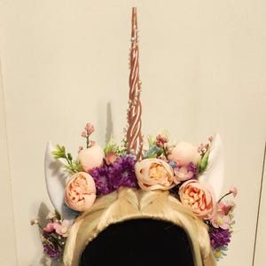 May include: A whimsical unicorn headpiece featuring a rose gold horn, white ears, and a floral crown with pink and purple flowers. The headpiece is set on a black mannequin head with blonde hair and long white eyelashes.