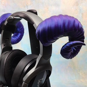 May include: Black over-ear headphones featuring two large, curved, purple ram-style horns attached to the headband. The horns have a shiny, satin-like texture with visible ridges, and the headphones include black ear cushions and a dark gray frame.