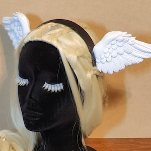 May include: A black headband with attached white angel wings. The wings are detailed with feather textures. The headband is worn on a mannequin head with blonde hair and long white eyelashes.
