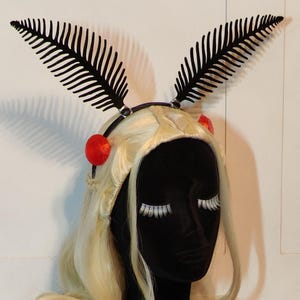 May include: A black headband featuring two fern-shaped decorations. The headband is decorated with red spheres and is displayed on a black mannequin head with a blonde wig and white eyelashes. The wig has long, flowing hair.