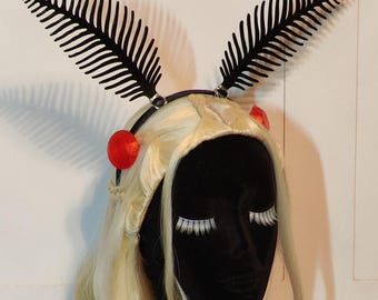 Könnte beinhalten: Ein schwarzes Stirnband mit zwei stilisierten Farn-Akzenten. Das Stirnband ist mit roten Kugeln verziert und befindet sich auf einem schwarzen Schaufensterkopf mit einer blonden Perücke und weißen Wimpern. Die Perücke hat lange, fließende Locken.