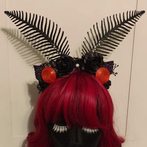 May include: A black headpiece with fern-like accents, black roses, and orange orbs. The headpiece is adorned with small pearls and sits atop a mannequin head with red hair and white eyelashes. The headpiece is a unique accessory.
