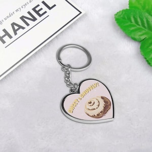 May include: A silver heart-shaped keychain with a pink background and the text "Sweet & Snuggly?" in yellow. A cartoon cinnamon roll is pictured on the keychain.