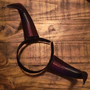 K&ouml;nnte beinhalten: Ein dunkelburgunderfarbenes Hexenhut-Stirnband mit gebogener Krempe und spitzer Spitze. Das Stirnband ist schwarz und verbindet sich mit der Hutbasis. Der Hut besteht aus gl&auml;nzendem Material und ist vor einem Holzgrund platziert.