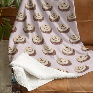 May include: A soft, plush throw blanket with a white background and a repeating pattern of brown cinnamon rolls. The blanket is folded and draped over a brown leather couch.