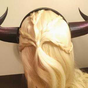 K&ouml;nnte beinhalten: Ein schwarzes und lila geh&ouml;rntes Stirnband mit geflochtenem Design. Die H&ouml;rner sind nach oben und au&szlig;en gebogen. Das Stirnband wird auf blondem Haar getragen, das mit Z&ouml;pfen versehen ist.