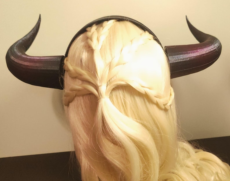 K&ouml;nnte beinhalten: Ein schwarzes und lila geh&ouml;rntes Stirnband mit geflochtenem Design. Die H&ouml;rner sind nach oben und au&szlig;en gebogen. Das Stirnband wird auf blondem Haar getragen, das mit Z&ouml;pfen versehen ist.