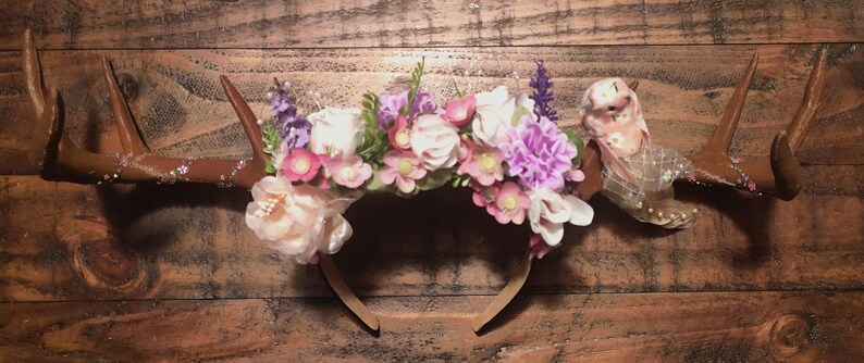 May include: A floral headband with antlers and a delicate tulle veil. The headband is adorned with pink, white, and purple flowers.