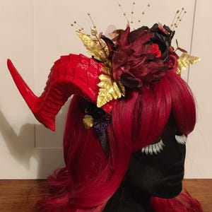 May include: A red horned headpiece with gold and burgundy floral accents. The headpiece features a large, textured red horn, gold leaves, and burgundy flowers. The headpiece is set against a backdrop of red hair.
