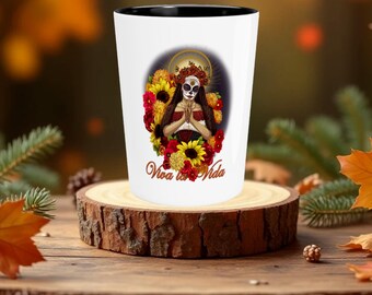 May include: White ceramic mug with a black rim. The mug features a colorful illustration of a woman with a sugar skull face surrounded by flowers. The text "Viva la Vida" is printed on the mug.