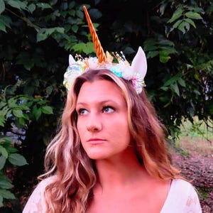 May include: A unicorn headband featuring a tall, spiraled metallic gold horn centered between two white ears. The base is adorned with iridescent, multi-colored floral accents, small white flowers, and delicate gold-toned wire embellishments.