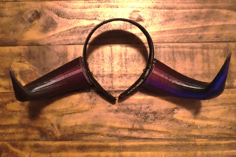 K&ouml;nnte beinhalten: Ein schwarzes Stirnband mit zwei gebogenen H&ouml;rnern. Die H&ouml;rner gehen von Schwarz &uuml;ber Lila zu Blau &uuml;ber. Die H&ouml;rner bestehen aus einem 3D-gedruckten Material und liegen auf einer Holzoberfl&auml;che.