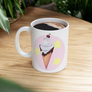 Könnte beinhalten: Eine weiße Keramiktasse mit einem rosa und gelben Design, das eine weiße Schlange in einem Waffelhörnchen mit einer Kirsche oben zeigt. Die Tasse ist mit Kaffee gefüllt.