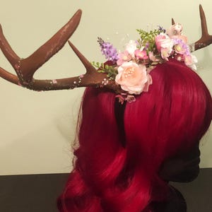 May include: A red wig with a floral crown featuring pink and purple flowers, and a pair of brown antlers with glitter accents.