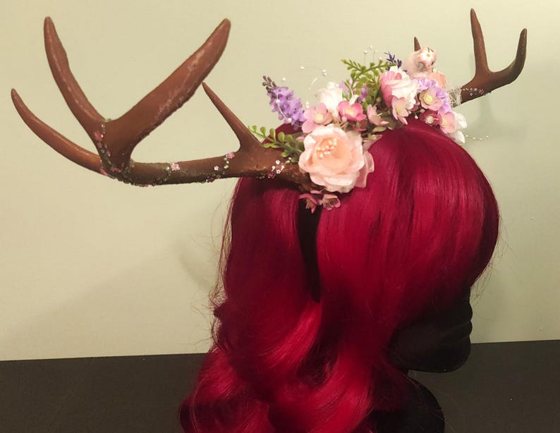 May include: A red wig with a floral crown featuring pink and purple flowers, and a pair of brown antlers with glitter accents.