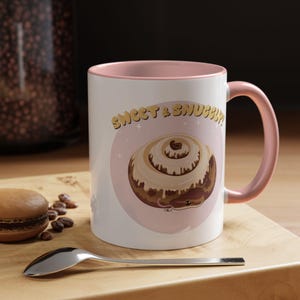 May include: White ceramic mug with a pink interior and handle. The mug features a cinnamon roll design with a snake's head and the text "Sweet & Snuggly" in gold lettering. The mug is on a wooden surface.