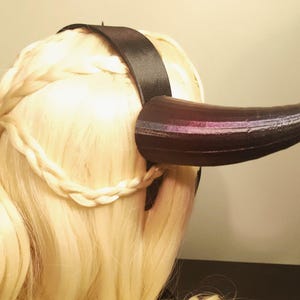 K&ouml;nnte beinhalten: Ein schwarzes Horn-Stirnband mit einem schwarzen Satinband. Das Horn ist dunkelbraun mit lila und silberfarbenen Akzenten. Das Haar ist blond und zu einem Zopf geflochten.