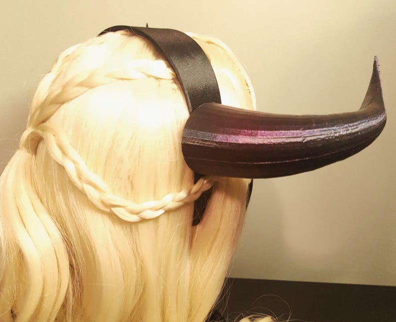 K&ouml;nnte beinhalten: Ein schwarzes Horn-Stirnband mit einem schwarzen Satinband. Das Horn ist dunkelbraun mit lila und silberfarbenen Akzenten. Das Haar ist blond und zu einem Zopf geflochten.