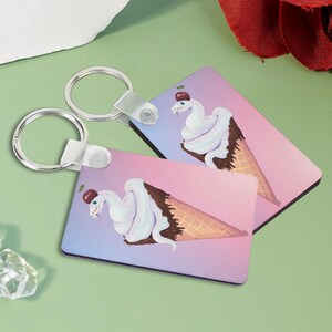 Könnte beinhalten: Zwei Schlüsselanhänger mit einer weißen Schlange, die um einen Eiswaffel herumgewickelt ist. Die Schlange hat eine rote Kirsche auf dem Kopf. Der Eiswaffel befindet sich auf einem rosa und blauen Hintergrund.