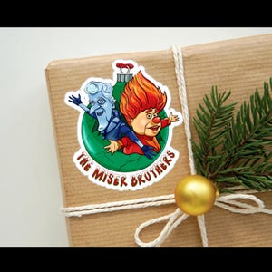May include: A green Christmas ornament with a white and blue character and a red and orange character inside. The ornament has the text "The Miser Brothers" around the outside.