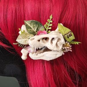 May include: A unique hair accessory featuring a dinosaur skull, accented with faux flowers and greenery. The skull is cream-colored, with sharp teeth and detailed bone structure. The floral elements include pink and white blossoms and various green leaves.