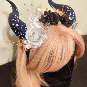 May include: A dark blue and gold horned headband with white and black flowers. The horns are decorated with gold stars and the flowers are made of fabric.