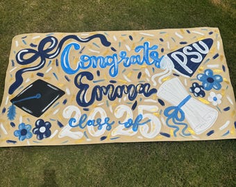 Op de afbeelding: Een handgeschilderde afstudeerbanner met de woorden "Congrats Emma Class of 25" in blauw en wit. De banner bevat een afstudeerhoed, diploma en bloemen. De achtergrond is beige met gouden accenten.