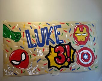 Può includere: Un banner dipinto a mano a tema supereroi. Il banner presenta il nome "LUKE is 3!" in lettere blu e rosse, insieme a illustrazioni di un pugno verde di Hulk, Spider-Man, Iron Man e lo scudo di Capitan America. Lo sfondo è decorato con coriandoli colorati.
