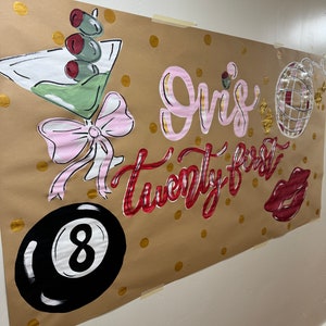 Op de afbeelding: Een bruine papieren banner met gouden en witte stippen. Op de banner staat in roze en rode cursieve letters "Griz's twenty-first". Er is een roze strik, een martini-glas met olijven, een zwart-witte 8-ball en een rode discobal.