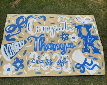 Op de afbeelding: Een handgeschilderd afstudeerbord met blauwe en witte letters op een bruine achtergrond. Het bord zegt "Gefeliciteerd Thanya klas van 2023" met het logo van de Universiteit van Kentucky en bloemmotieven.