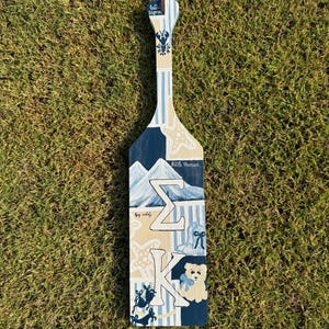 Puede incluir: Una paleta de madera con un lazo de cuero marrón para colgar. La paleta está pintada con rayas azules y blancas y presenta una langosta, una estrella de mar, montañas y las letras griegas Sigma y Kappa, junto con un oso de peluche.