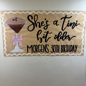 May include: A brown paper banner with white scalloped edges. The banner has black lettering that reads "She's a tiny bit older" and "Morgan's 30th Birthday". A drawing of a cocktail glass with a pink bow is on the left side of the banner.