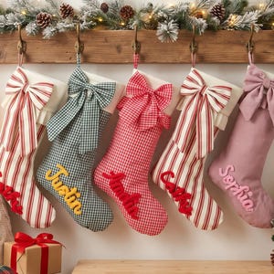 Puede incluir: Cinco calcetines navideños colgantes con nombres personalizados en letra cursiva. Los calcetines presentan patrones de rayas rojas y blancas, cuadros vichy verdes y blancos, cuadros rojos y blancos y una tela rosa empolvado, cada uno con un gran lazo.