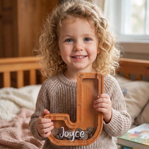 Può includere: Un salvadanaio in legno a forma di lettera J con un pannello frontale in acrilico trasparente. Il salvadanaio è riempito con varie monete metalliche e presenta il nome Joyce scritto in corsivo bianco sulla superficie del pannello trasparente.