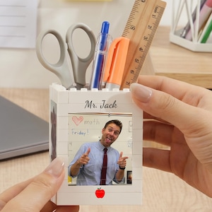 Puede incluir: Un organizador de escritorio rectangular blanco construido con bloques, que presenta una fotografía impresa de un hombre señalando a la cámara, el texto "Mr. Jack" y un pequeño icono de manzana roja. Contiene tijeras grises, un bolígrafo azul, un marcador naranja y una regla de madera.