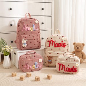 May include: Two corduroy backpacks and two matching lunch bags featuring a strawberry pattern. One set is dusty pink with the name Amelia in pastel letters, the other is beige with the name Mavis in red letters. Each item has a small flower and plush toy.