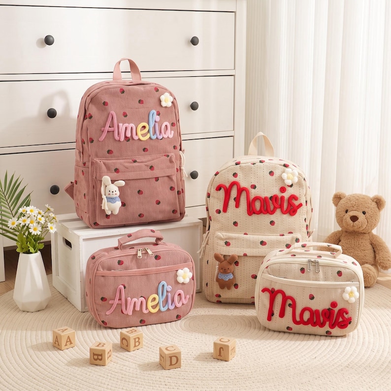 May include: Two corduroy backpacks and two matching lunch bags featuring a strawberry pattern. One set is dusty pink with the name Amelia in pastel letters, the other is beige with the name Mavis in red letters. Each item has a small flower and plush toy.