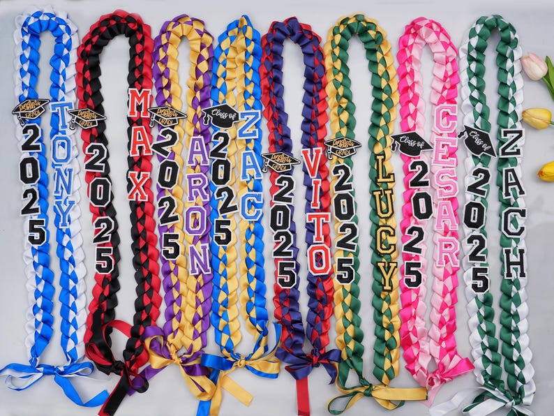 May include: Nine graduation leis with different colored ribbons and personalized patches. The patches include names like 'Tony', 'Max', 'Aaron', 'Zach', 'Vito', 'Lucy', 'Cesar', and 'Zach' with the year '2025' on each. The leis are made with blue, white, red, black, purple, yellow, green, pink, and white ribbons.