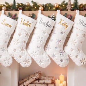 May include: Five white faux fur Christmas stockings hanging in a row, each featuring embroidered snowflake patterns and personalized names in gold or silver script: Luna, Violet, Barbie, Katty, and Milo. The stockings have plush cuffs.